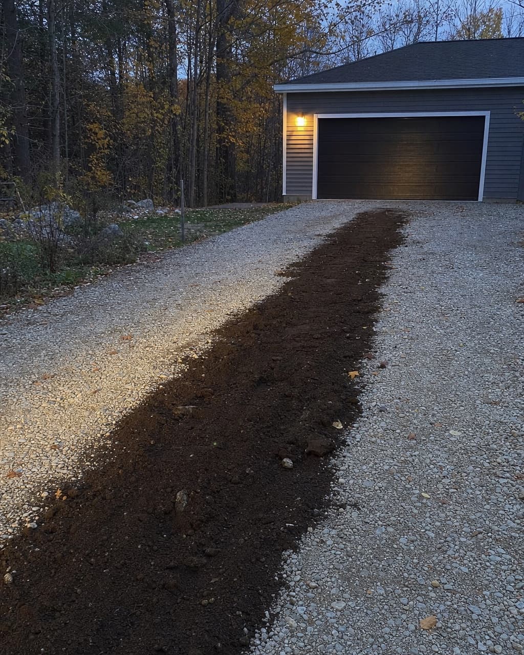 Une tranchée fraîchement rebouchée traversant une allée de gravier vers un garage détaché, borne allumée au mur